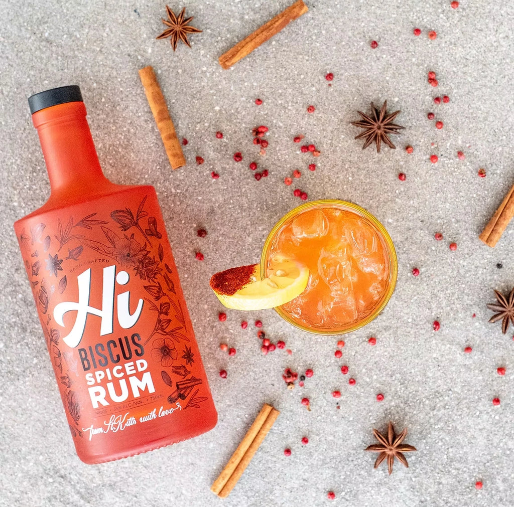 Hi Biscus Spiced Rum bottle in a flat-lay with a Spiced Rum Punch cocktail, cinnamon sticks, star anise and pink peppercorns