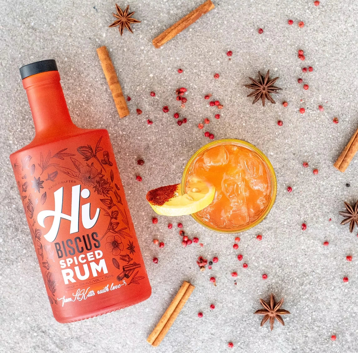 Hi Biscus Spiced Rum bottle in a flat-lay with a Spiced Rum Punch cocktail, cinnamon sticks, star anise and pink peppercorns